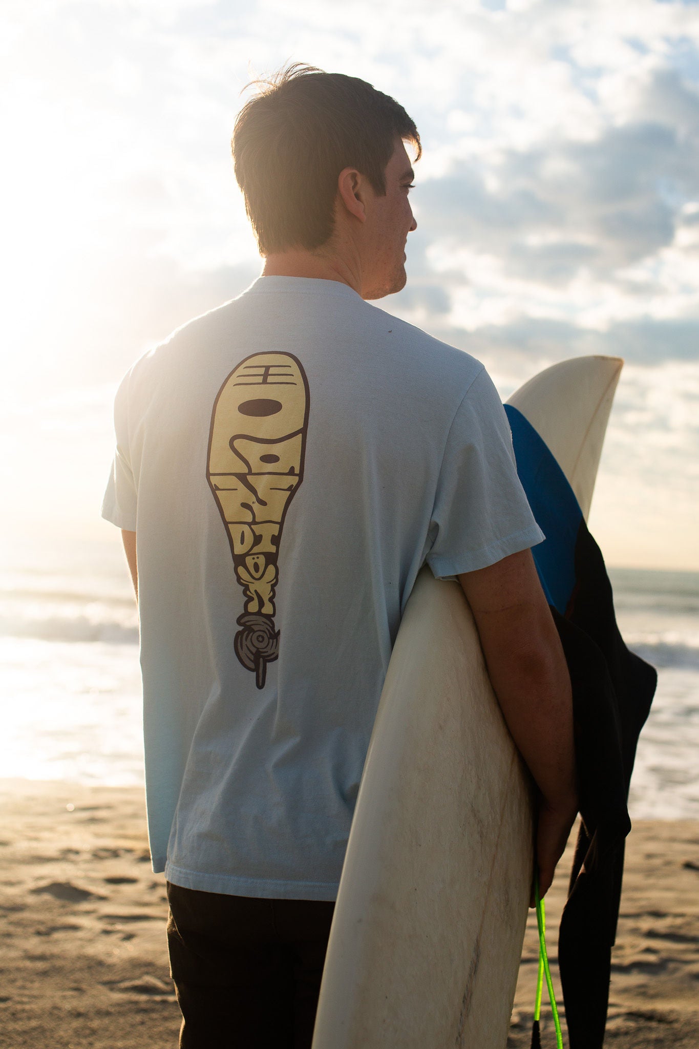 outboard light blue tee shirt on man carrying a surfboard on the beach